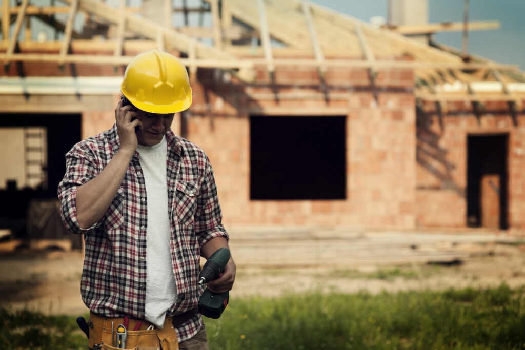 A man in a hard hat talking on a cellphone in front of a house under construction